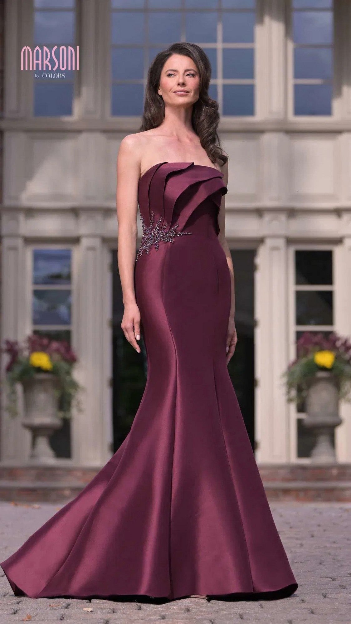 A woman wearing the Marsoni MV1308 long formal dress, a strapless mermaid gown with decorative pleats and beaded applique, stands outdoors in front of a large building.